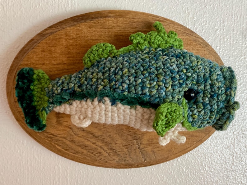 May include: A handmade crocheted fish, in shades of blue and green, mounted on an oval wooden plaque. The fish has detailed scales and fins, with a dark eye. A unique, handcrafted piece for the home.