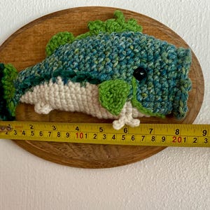May include: A handmade crochet fish mounted on a wooden plaque. The fish is primarily blue and green, with white and green accents. The fish measures approximately 23 cm long, as indicated by the measuring tape.