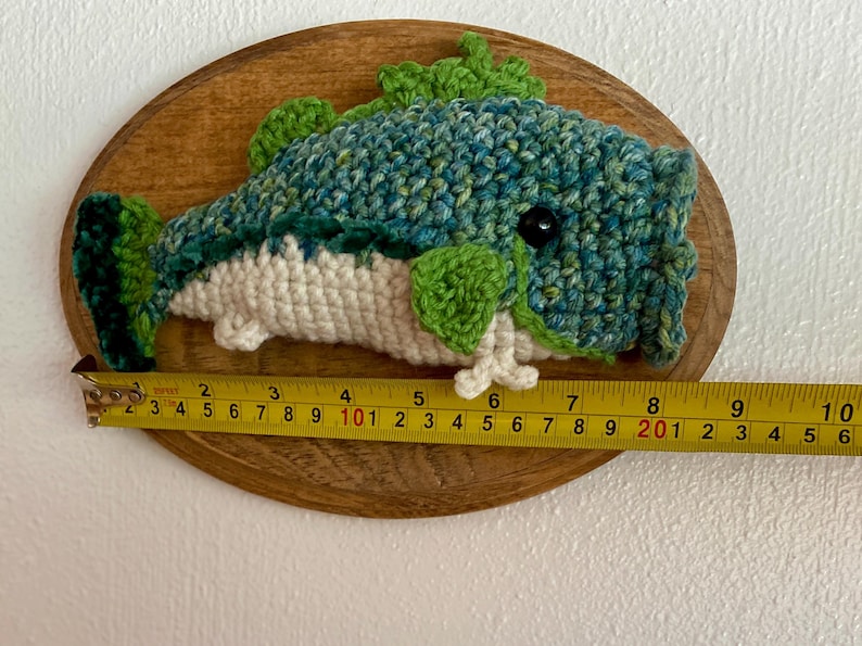 May include: A handmade crochet fish mounted on a wooden plaque. The fish is primarily blue and green, with white and green accents. The fish measures approximately 23 cm long, as indicated by the measuring tape.