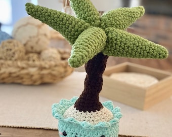 May include: A crocheted palm tree toy with a dark brown trunk and green fronds, planted in a light blue crocheted pot. The pot features a stitched smiling face with black eyes, a curved mouth, and small pink cheeks, with a scalloped top edge.