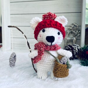 May include: A handmade white crochet polar bear wearing a red hat with a pom-pom, a red and white scarf, and a brown bag. The bear is holding a fishing rod with a gray fish attached. A charming winter-themed decoration.