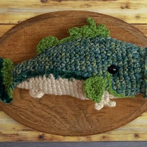 May include: A handcrafted crochet fish in shades of blue, green, and cream, mounted on an oval wooden plaque. The fish features a textured design with a black eye and green fins. The wooden plaque is approximately 30 cm long.