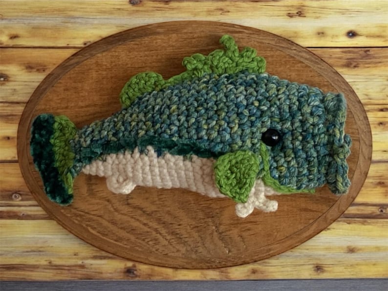 May include: A handcrafted crochet fish in shades of blue, green, and cream, mounted on an oval wooden plaque. The fish features a textured design with a black eye and green fins. The wooden plaque is approximately 30 cm long.