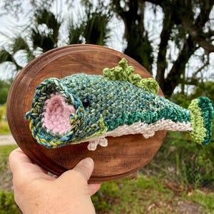 May include: A crochet fish mounted on a round wooden plaque. The fish is made with green, blue, and white yarn, with a pink mouth and black eyes. The wooden plaque is brown, creating a rustic aesthetic. The fish is a decorative item.