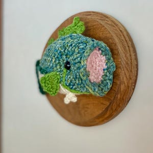May include: A crocheted fish wall hanging in shades of teal and green, with pink detailing and a black eye. The fish is mounted on a circular wooden plaque, offering a unique piece of home decor for any room.