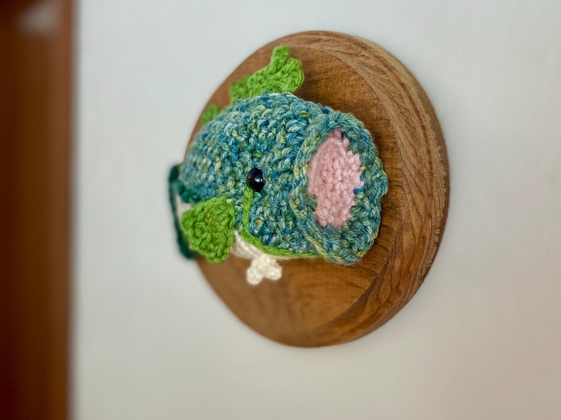 May include: A crocheted fish wall hanging in shades of teal and green, with pink detailing and a black eye. The fish is mounted on a circular wooden plaque, offering a unique piece of home decor for any room.