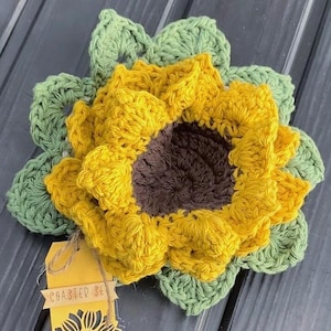 May include: A handmade crochet sunflower coaster set. The coaster features a yellow flower with a brown center and green leaves. A small tag attached to the coaster reads "Coaster Set".