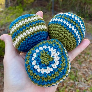 May include: Three handmade crochet balls in shades of blue, green, and cream. The balls are held in a hand, showcasing the textured stitch work. The balls are designed with stripes and circular patterns.