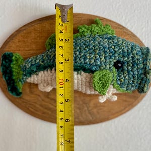 May include: A handmade crochet fish wall hanging, approximately 18 cm long, mounted on an oval wooden plaque. The fish features shades of blue and green yarn with a white belly and green fins. A yellow tape measure is next to the fish.