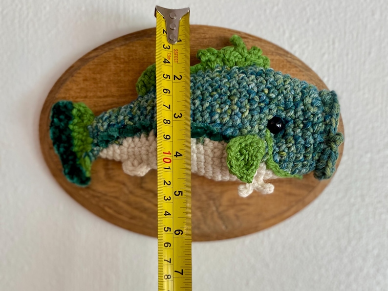 May include: A handmade crochet fish wall hanging, approximately 18 cm long, mounted on an oval wooden plaque. The fish features shades of blue and green yarn with a white belly and green fins. A yellow tape measure is next to the fish.