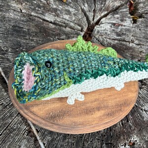 May include: A crocheted fish sculpture in shades of green, blue, and white, mounted on a round wooden plaque. The fish has an open mouth with pink detailing and is displayed on a natural wood surface.