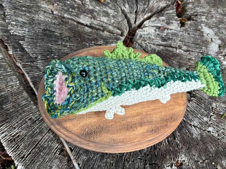May include: A crocheted fish sculpture in shades of green, blue, and white, mounted on a round wooden plaque. The fish has an open mouth with pink detailing and is displayed on a natural wood surface.