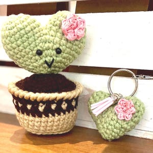Peut inclure: Cactus en forme de cœur crocheté, avec une fleur rose et un visage souriant, dans un pot marron et beige. Un porte-clés assorti en forme de cœur avec un pompon et une fleur roses est également présenté.