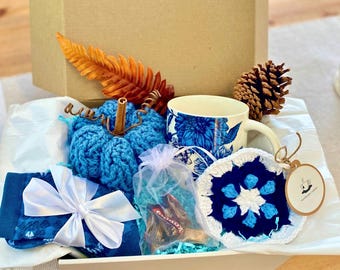 May include: A gift box filled with a blue crocheted pumpkin, a floral-patterned mug, a white-bowed item, a crocheted coaster, and a pinecone. The box includes a decorative orange leaf and a small bag of chocolates.