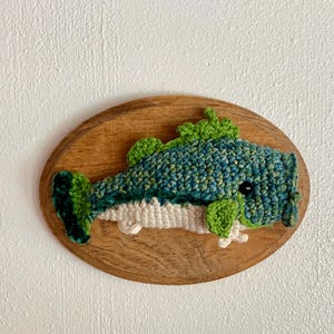 May include: A handmade crochet fish wall hanging. The fish is predominantly blue and green, with white detailing, mounted on an oval wooden plaque. The fish has a detailed, textured appearance, with a whimsical design.