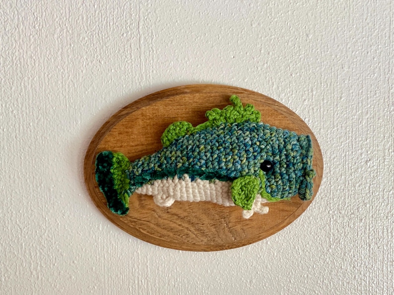 May include: A handmade crochet fish wall hanging. The fish is predominantly blue and green, with white detailing, mounted on an oval wooden plaque. The fish has a detailed, textured appearance, with a whimsical design.