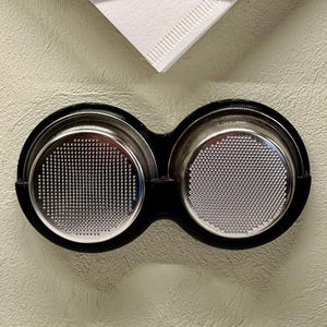 May include: Two silver-colored espresso portafilters with a black holder, mounted on a light beige wall. The portafilters have a fine mesh design.