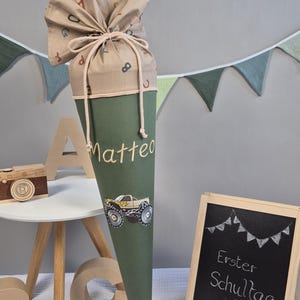 Könnte beinhalten: Eine grüne Schultüte mit dem Namen "Matteo" und einer Monster-Truck-Grafik. Die Tüte ist mit einem Kordelzug gebunden und hat einen gemusterten Deckel. Eine kleine Tafel mit dem Text "Erster Schultag" ist ebenfalls zu sehen.