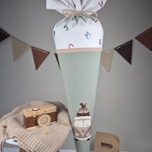 Könnte beinhalten: Eine große, mintgrüne, kegelförmige Schultüte mit einem weißen Oberteil, das mit bunten Buchstaben verziert ist. Die Tüte zeigt eine gestickte Illustration eines Oldtimer-Vans mit Gepäck und einer Katze. Eine Holzspielzeugkamera und ein kleiner Hocker sind ebenfalls im Bild.