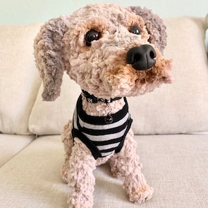 May include: A handmade plush dog with a black nose, dark eyes, and a black and white striped shirt. The dog has a black collar with a small charm. The dog is sitting on a light-coloured couch.