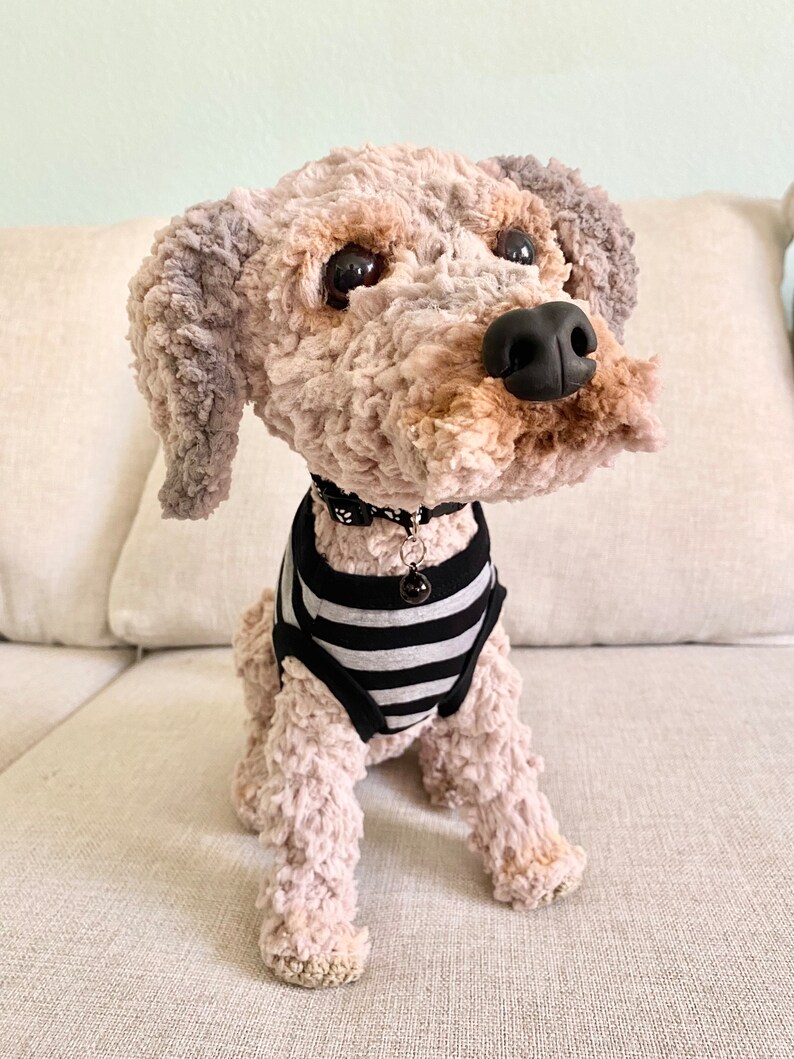 May include: A handmade plush dog with a black nose, dark eyes, and a black and white striped shirt. The dog has a black collar with a small charm. The dog is sitting on a light-coloured couch.