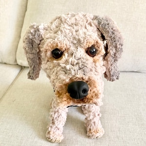 May include: A soft dog toy with a textured, cream-coloured body, brown and grey accents, and a black nose. The dog has large, dark eyes and floppy ears. The toy is designed to resemble a real dog and is positioned upright.