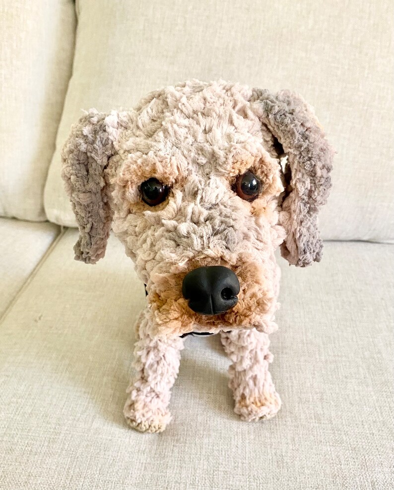 May include: A soft dog toy with a textured, cream-coloured body, brown and grey accents, and a black nose. The dog has large, dark eyes and floppy ears. The toy is designed to resemble a real dog and is positioned upright.