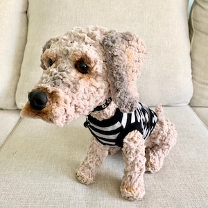 May include: A crocheted dog toy wearing a black and white striped top. The dog has a black nose, brown eyes, and a black collar. It is sitting on a beige sofa.