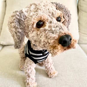 May include: A soft toy dog with a curly, textured coat in beige, grey, and brown. The dog wears a black and white striped shirt and has a black nose and dark eyes. A small bell hangs from its collar.