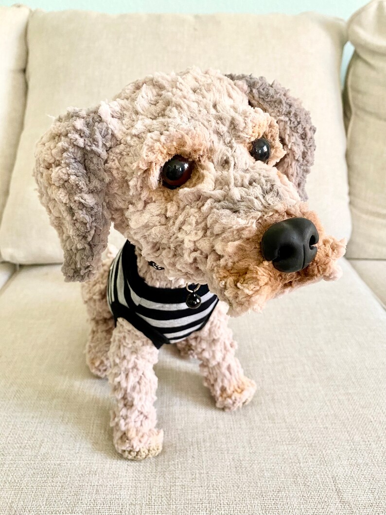 May include: A soft toy dog with a curly, textured coat in beige, grey, and brown. The dog wears a black and white striped shirt and has a black nose and dark eyes. A small bell hangs from its collar.