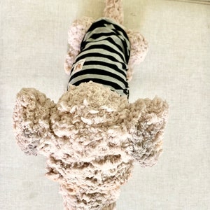 May include: A soft dog toy with a light brown, textured body and a black and grey striped shirt. The toy has a black nose and floppy ears. The dog is lying on a light-coloured surface.