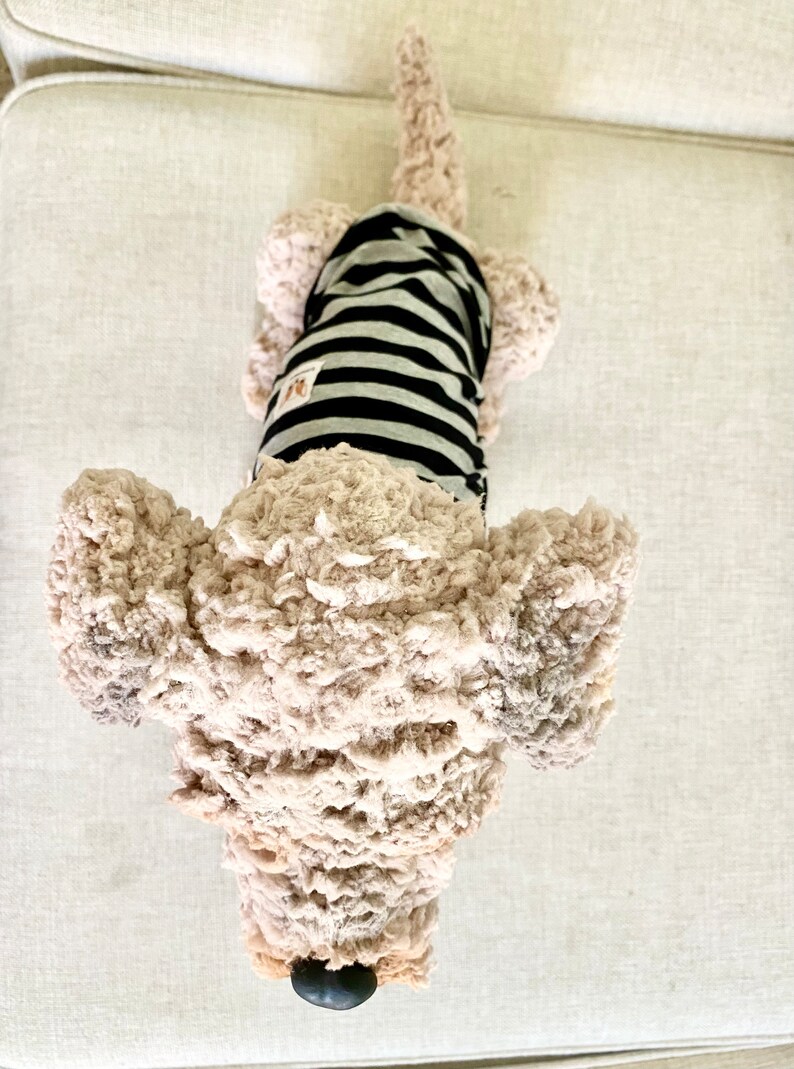 May include: A soft dog toy with a light brown, textured body and a black and grey striped shirt. The toy has a black nose and floppy ears. The dog is lying on a light-coloured surface.