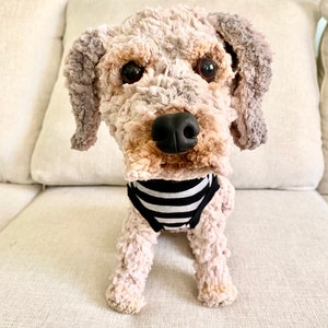 May include: A plush dog toy with a light brown and gray textured body, black eyes, and a black nose. The dog is wearing a black and white striped garment. The toy is sitting on a beige couch.