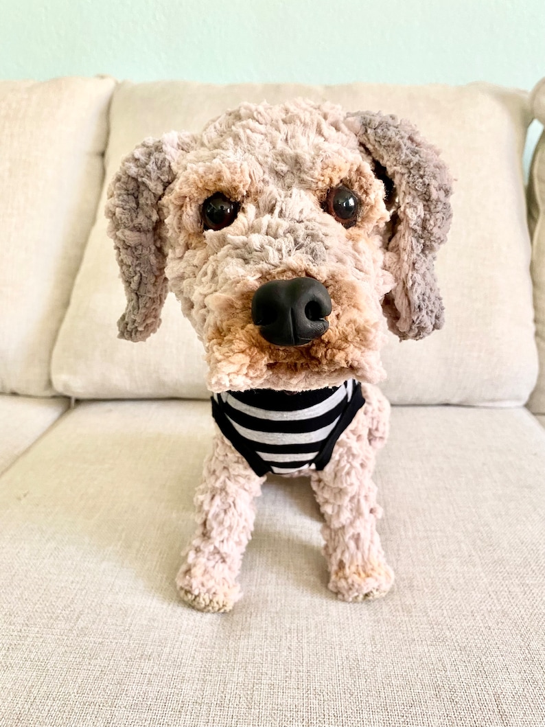 May include: A plush dog toy with a light brown and grey textured body, black eyes, and a black nose. The dog is wearing a black and white striped garment. The toy is sitting on a beige sofa.