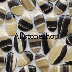 May include: A collection of 20 cabochon gemstones with black and white stripes. The stones are oval and teardrop shaped. Allstoneshop is printed on the image.