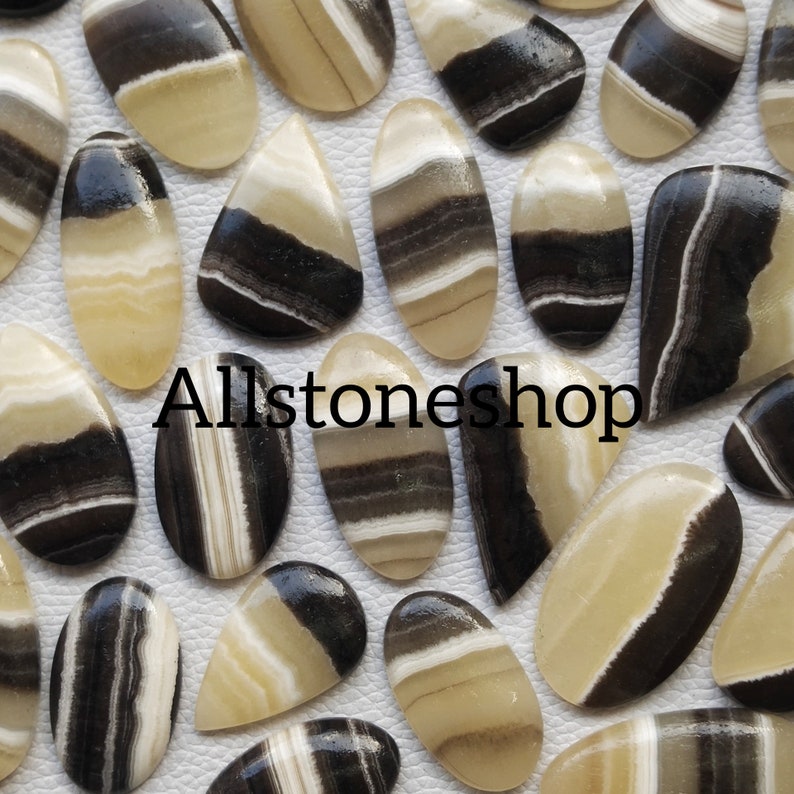 May include: A collection of 20 cabochon gemstones with black and white stripes. The stones are oval and teardrop shaped. Allstoneshop is printed on the image.