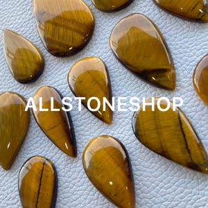May include: A collection of 10 teardrop-shaped tiger's eye gemstone cabochons. The stones are a rich golden brown color with a chatoyant effect. The stones are all different sizes and shapes, but they are all polished and smooth.