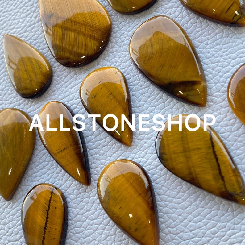 May include: A collection of 10 teardrop-shaped tiger's eye gemstone cabochons. The stones are a rich golden brown color with a chatoyant effect. The stones are all different sizes and shapes, but they are all polished and smooth.