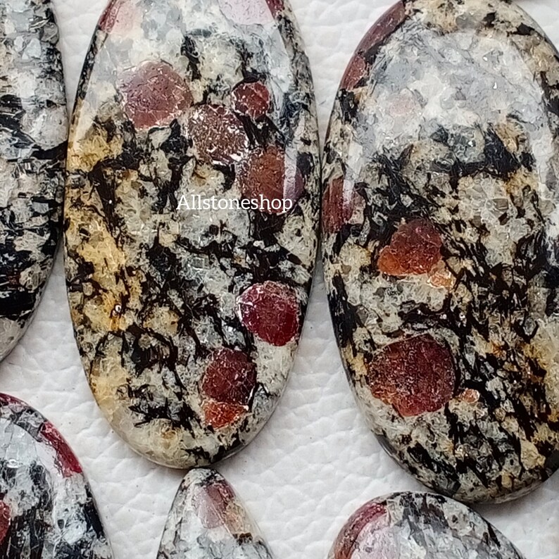 May include: Close-up of several oval gemstones. The stones have a speckled appearance with cream, black, and red colours. The gemstones are polished and have a smooth surface. The text "Allstoneshop" is visible.
