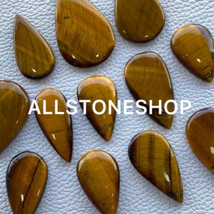 May include: A collection of ten teardrop-shaped tiger's eye gemstone cabochons. The stones are a mix of golden brown and black with a shimmering, iridescent effect. The stones are arranged on a white surface.