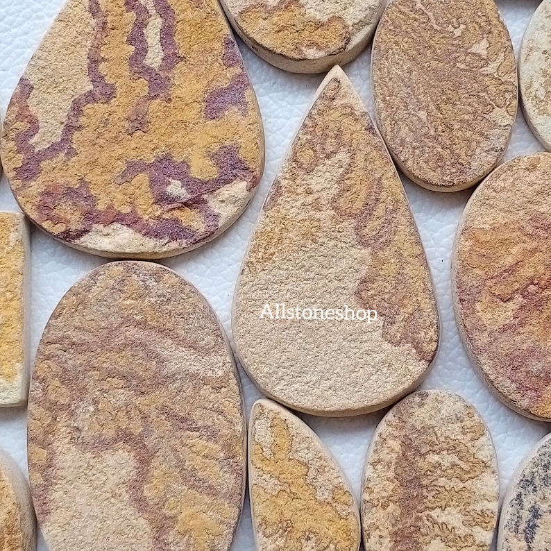 May include: A collection of natural stone cabochons in various shapes and sizes. The stones are a mix of brown, tan, and red with a unique pattern. Allstoneshop is printed on one of the stones.