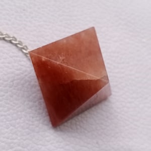 Può includere: Un ciondolo a forma di piramide di cristallo rosso su una catena d'argento. La piramide è sfaccettata e ha una finitura liscia e lucida.