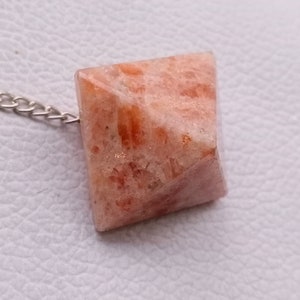 Może przedstawiać: Wisiorek w kształcie piramidy ze słonecznego kamienia z srebrnym łańcuszkiem. Słoneczny kamień jest jasno pomarańczowy z białym i pomarańczowym wzorem.