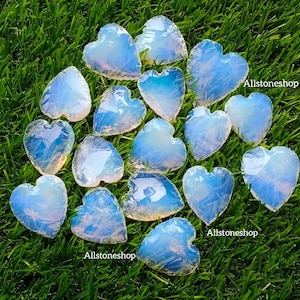 Peut inclure: Une collection de 14 pierres d'opale en forme de cœur bleu irisé sur une surface herbeuse verte. Les pierres sont disposées de manière aléatoire. Le texte "Allstoneshop" apparaît deux fois sur l'image.