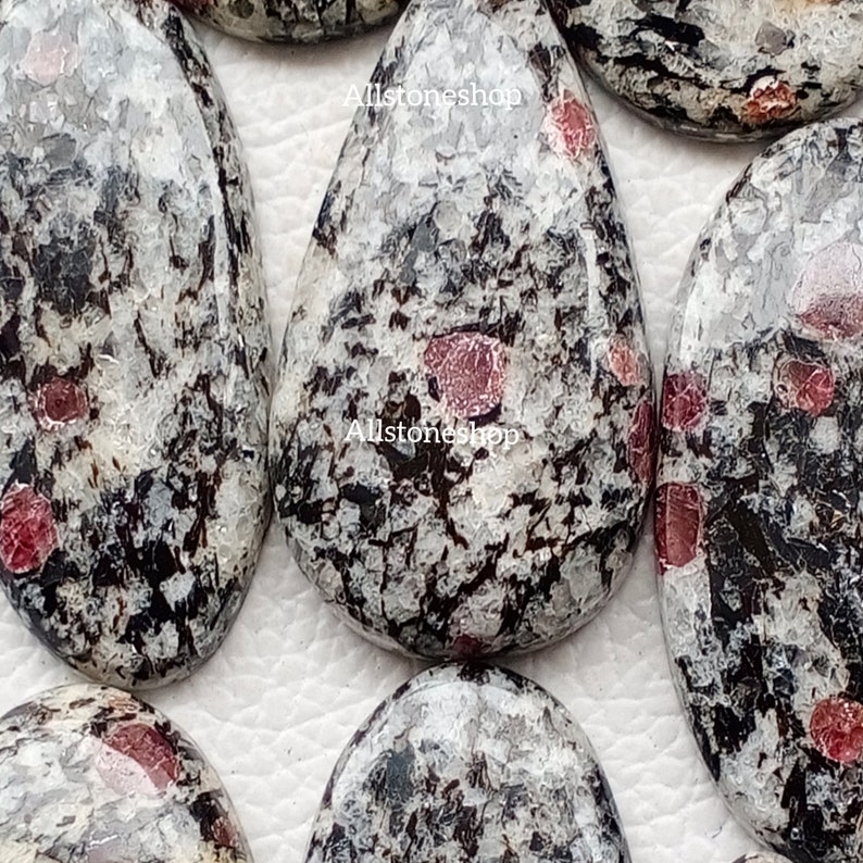May include: Close-up of several polished stones with a speckled pattern. The stones are primarily white and grey, with black veining and small, red inclusions. The stones have a smooth, glossy finish and are teardrop-shaped.
