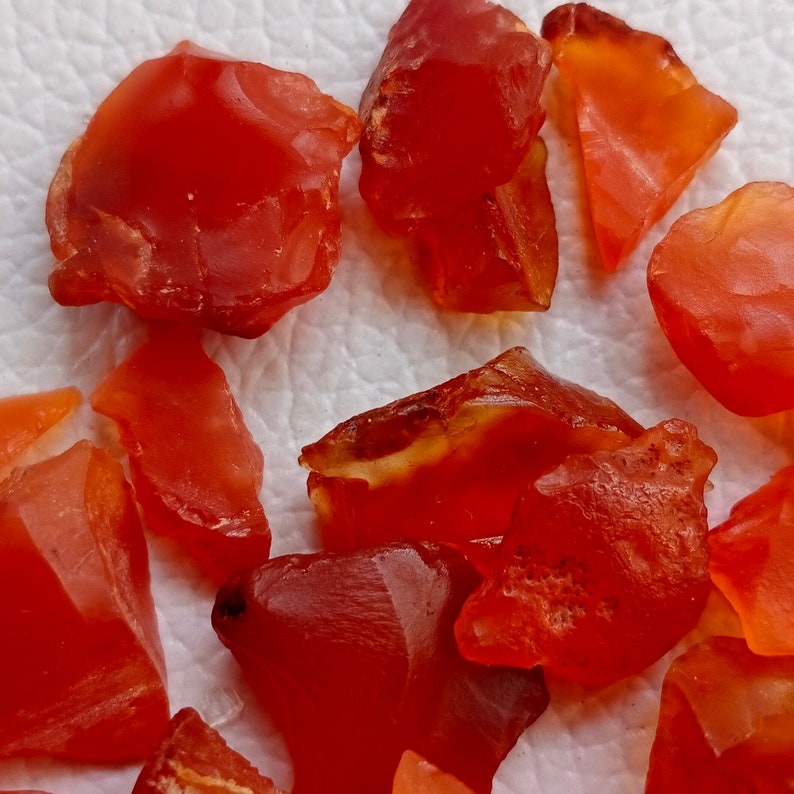 Puede incluir: Una colecci&oacute;n de piedras preciosas de cornalina roja, cada una con una forma y textura &uacute;nicas. Las piedras est&aacute;n dispuestas sobre una superficie blanca.