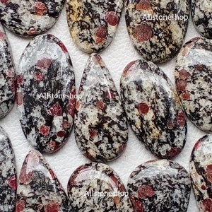 May include: A selection of polished gemstones with a speckled design. The stones feature a mix of black, white, and red hues. The gemstones are oval and teardrop shaped. The text "Allstoneshop" is visible on some of the stones.