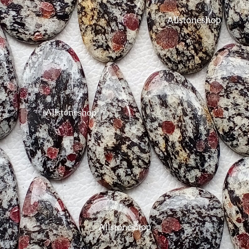 May include: A selection of polished gemstones with a speckled design. The stones feature a mix of black, white, and red hues. The gemstones are oval and teardrop shaped. The text "Allstoneshop" is visible on some of the stones.