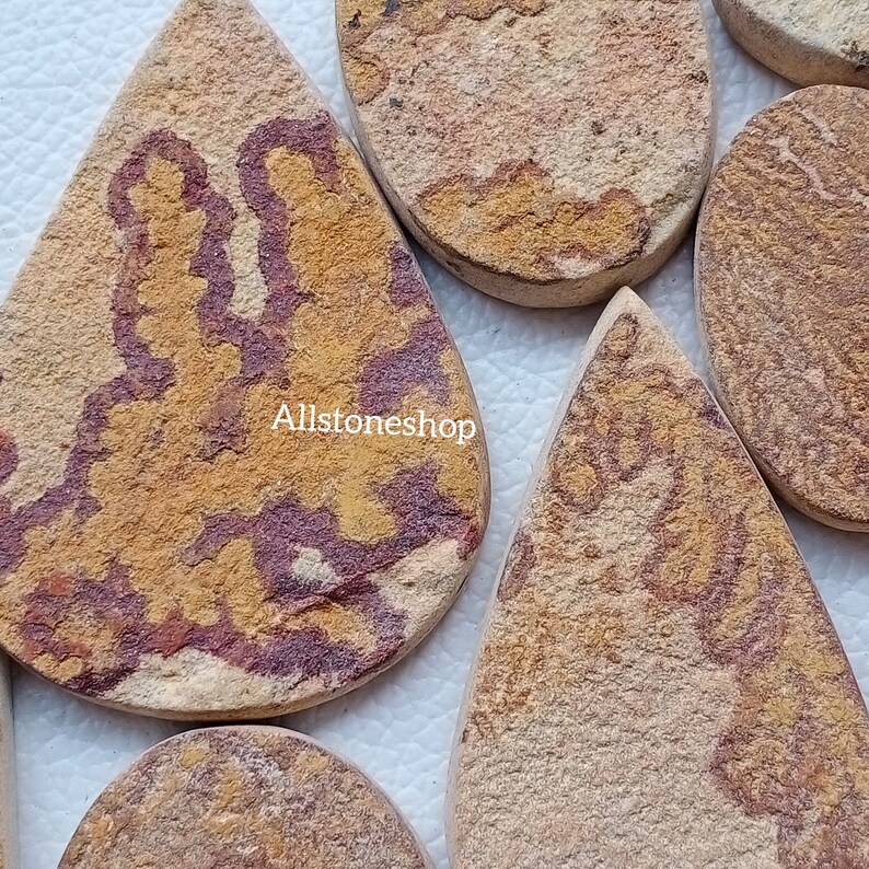 May include: A collection of six teardrop-shaped, polished sandstone cabochons with a variety of brown, yellow, and red patterns. The stones are all different sizes and shapes, but they all have a smooth, polished finish.