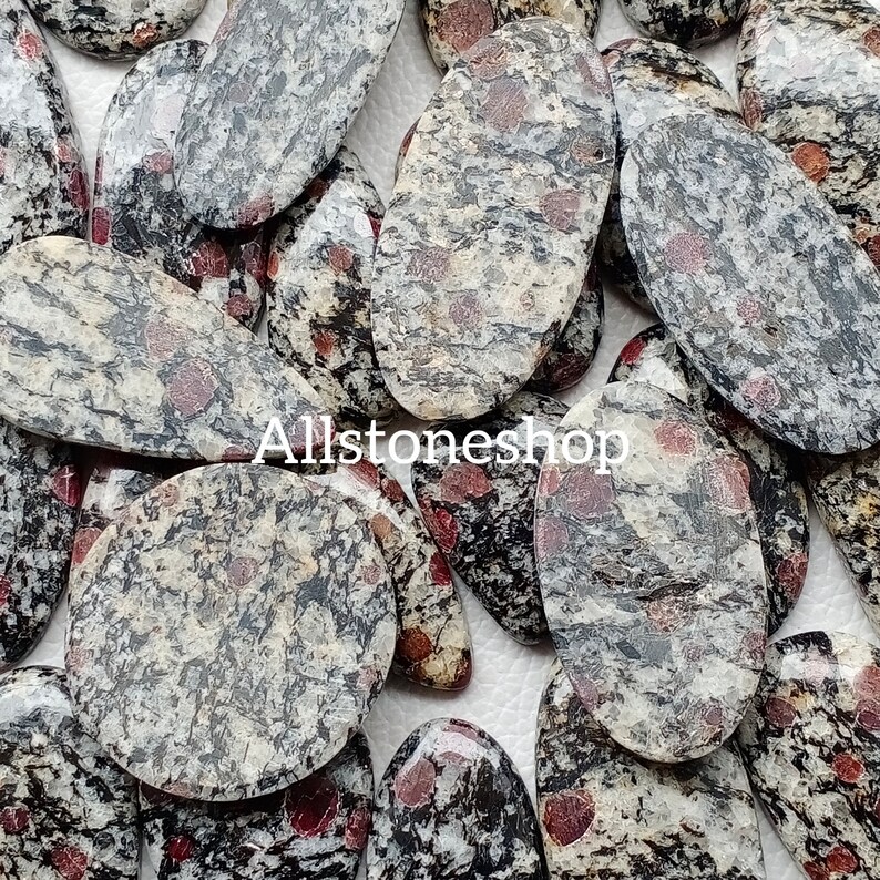 May include: A selection of oval and rounded stones with a speckled pattern of grey, black, and red. The stones have a polished appearance and vary in size. The text "Allstoneshop" is visible on the stones.
